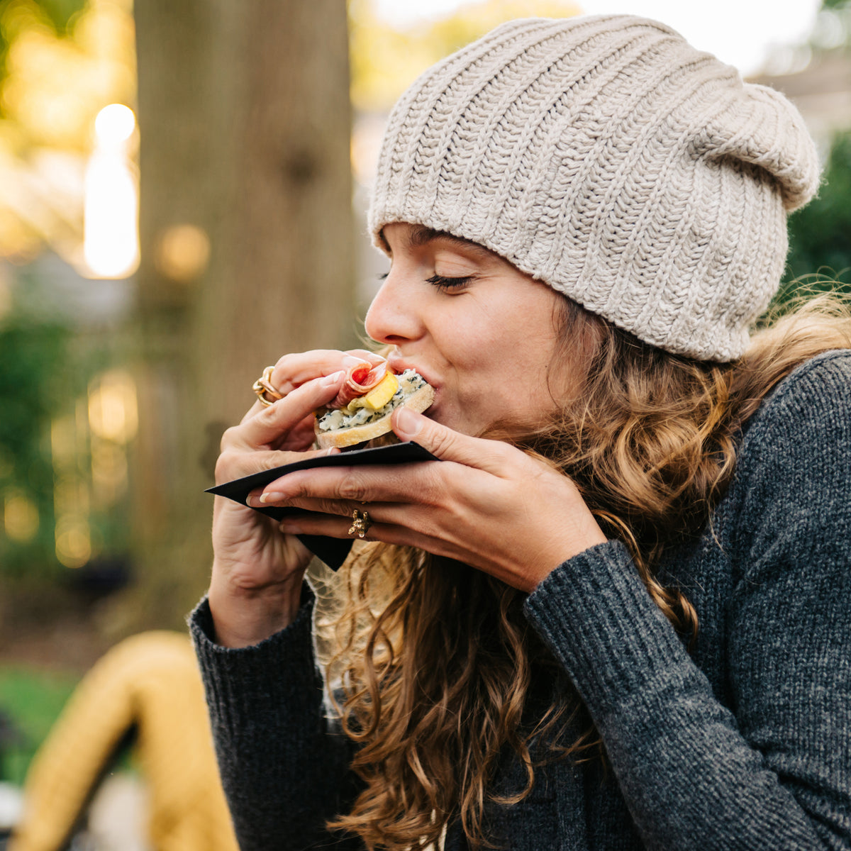 Lady eating a sandwich with a beanie on and appears to be in some form of woodland.