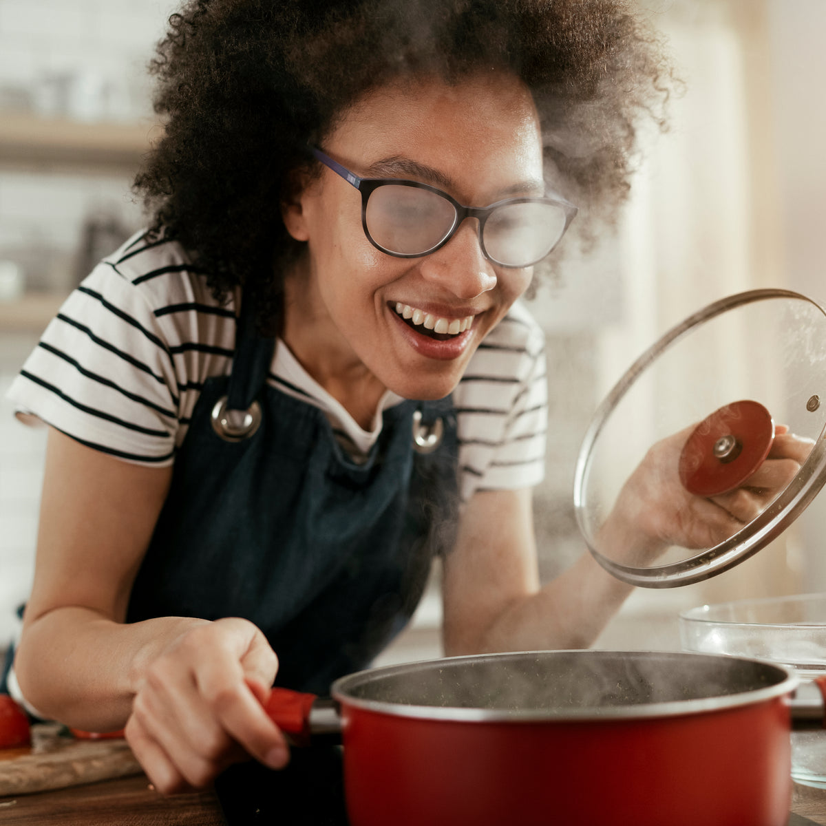 Lady who is smiling with steamed glasses as she looks into a pot on a stove.